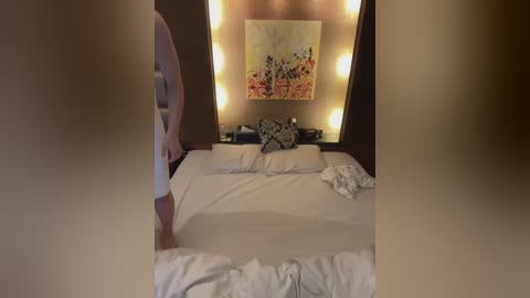 Media: Video of a modern, minimalist bedroom with a large bed, white linens, and a floral painting on the wall. Two wall-mounted lamps provide warm lighting.