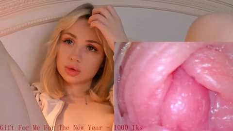 Video of a young blonde woman with fair skin, blue eyes, and shoulder-length hair, wearing a white jacket, reclining on a bed. Close-up inset shows her pink vaginal tissue.