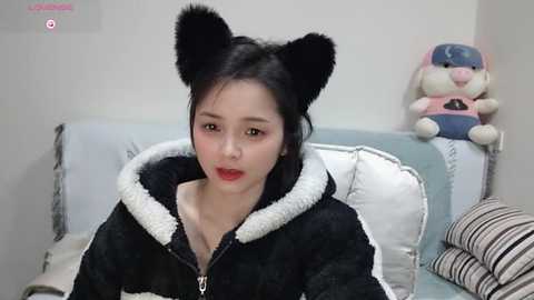 Media: Video of a young East Asian woman with fair skin, wearing a black hoodie with white fur trim, black cat ears, and red lipstick, sitting on a light blue couch.