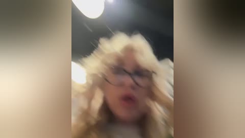 Media: A blurry video shows a blonde woman with curly hair and glasses, mouth open, in an indoor setting with a bright ceiling light.