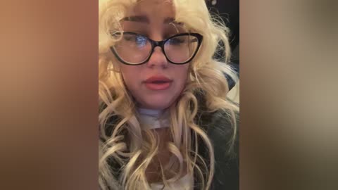 Media: Video of a young woman with long, wavy blonde hair, wearing large black-rimmed glasses and a black jacket. She has a fair complexion and full lips. The background is blurred, focusing on her face.