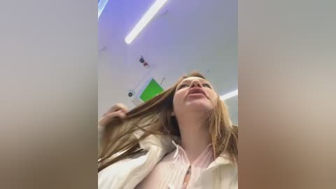 Media: Video of a young Caucasian woman with long, light brown hair, wearing a white shirt, tilted head, smiling, in a modern, brightly lit room with fluorescent lighting and green accents on the ceiling.