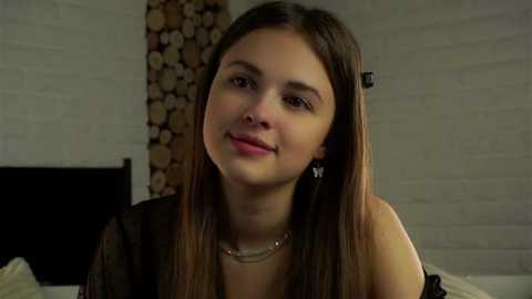 Video of a young woman with straight brown hair, fair skin, wearing a black top and silver necklace, smiling gently, standing in a rustic room with white brick walls and a wooden log stack in the background.