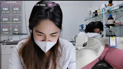 Video of an Asian woman in a lab coat, wearing a white face mask, examining a petri dish. Background shows lab equipment, bottles, and a digital display with health statistics.