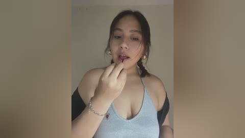 Media: Video of an Asian woman with medium skin tone, wearing a light blue tank top, biting her finger, against a plain beige wall.