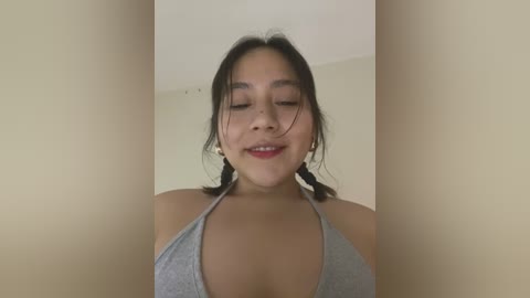 Media: Video of a young Asian woman with medium skin tone, wearing a grey halter top, smiling with her eyes closed, dark hair in braids, against a beige wall.