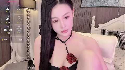 Media: A video of a fair-skinned East Asian woman with long, straight black hair, wearing a black halter top with red rose-shaped appliqu\u00e9s, sitting on a bed in a cozy, dimly lit bedroom.