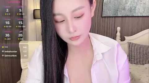 Media: Video of an East Asian woman with long black hair, wearing a white button-up shirt, sitting on a bed with a beige pillow, in a dimly lit room.