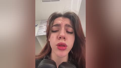 Media: Video of a young woman with fair skin, brown hair, and red lipstick, eyes closed, looking slightly shocked or surprised, in a modern office setting with white walls and a ceiling vent.