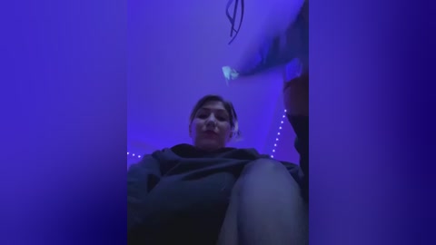 Media: Video of a woman with short dark hair, wearing a black hoodie, sitting in a dimly lit room with purple lighting.