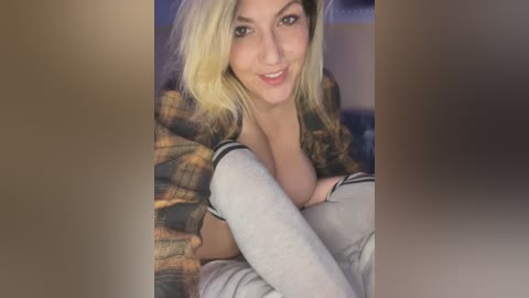 Media: A video of a smiling, light-skinned woman with blonde hair, wearing a plaid shirt and white leggings, seated on a bed.