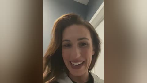 Media: A video of a smiling woman with light skin and long, wavy, auburn hair, wearing a white top, standing in a dimly lit room with grey walls and a white door.