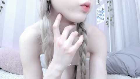 Media: Video of a pale-skinned woman with long platinum blonde hair, partially nude, kissing her wrist, in a softly lit bedroom with white curtains and plush pillows.