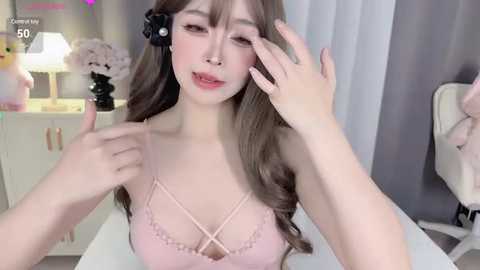 Media: A video of a young Asian woman with long brown hair, wearing a pink lace bra, adjusting her hair. She has fair skin and a slender build. The background features a white desk, a black lamp, and a pink flower arrangement.