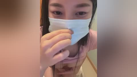 Media: Video of a young Asian woman with long black hair, wearing a pink shirt and a blue surgical mask, holding a piece of cloth over her nose, in an indoor setting.