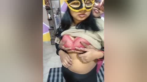 Media: A video captures a pregnant woman with long black hair, wearing a yellow mask and a beige top lifted to expose her large breasts. She holds her swollen belly with both hands. The background shows a colorful wall and a shelf with bottles.