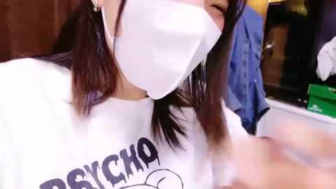 Media: Video of an Asian woman with dark hair, wearing a white mask and \"PSYCHO\" t-shirt, indoors, smiling.