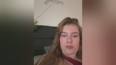 Media: Video of a young Caucasian woman with long, wavy brown hair, light skin, and a neutral expression, sitting in a dimly lit room with a green plant and a black leather couch in the background.