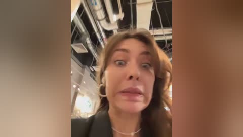 Media: Video of a shocked woman with wide eyes and open mouth in a busy, modern indoor setting with ceiling lights and hanging signs.