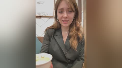 Media: Video of a young woman with light skin and long, wavy brown hair, wearing a dark blazer, sitting in a dimly lit diner booth, holding a white coffee cup.