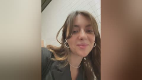 Media: A blurry video of a young woman with shoulder-length brown hair, wearing a black jacket, standing in a tiled hallway with beige walls. She has a subtle smile.