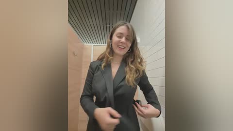 Media: Video of a smiling young woman with wavy brown hair, wearing a black blazer, standing in a narrow, beige-tiled hallway with white subway tiles and a black ceiling.