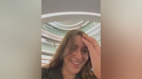 Media: Video of a young woman with long, wavy brown hair, wearing a black jacket, smiling and touching her forehead, set against a modern, circular, glass ceiling interior.