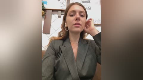 Media: Video of a young Caucasian woman with light skin, brown hair, wearing a grey blazer, earrings, and a necklace, sitting in an office, adjusting her ear.