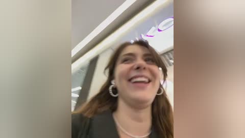 Media: Video of a smiling woman with long brown hair, wearing a black blazer, and silver hoop earrings, standing in a modern, brightly lit office space with glass walls and purple accents.