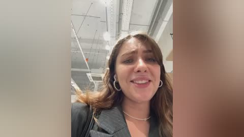 Media: A video of a young woman with long brown hair, wearing a dark blazer and pearl necklace, smiling in an office with white ceiling tiles and fluorescent lights.