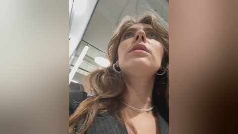 Media: Video of a young Caucasian woman with wavy brown hair and a fair complexion, wearing a black top and silver hoop earrings, looking upward with a neutral expression.