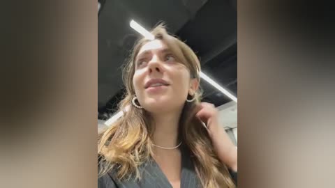 Media: Video of a young woman with long, wavy brown hair, wearing a grey shirt, looking upward with a thoughtful expression in a dimly lit room.