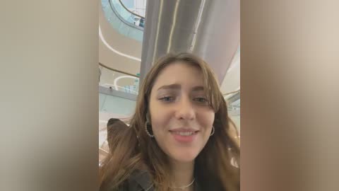 Media: Video of a young woman with light brown hair and fair skin, smiling, wearing a black top, standing in a modern, brightly lit shopping mall with glass railings and circular walkways in the background.