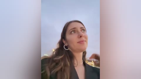 Media: Video of a young woman with long, wavy brown hair, wearing a black top and large silver hoop earrings, standing outdoors against a blurred, pale blue sky, with a soft, dreamy atmosphere.