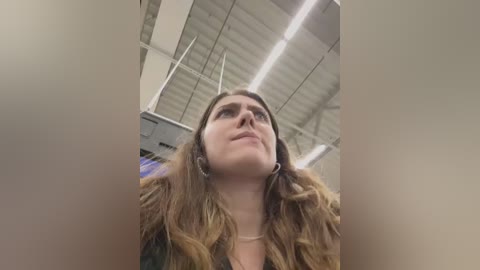 Media: Video of a Caucasian woman with wavy brown hair and fair skin, wearing a dark jacket, looking upward in a modern, industrial-style room with white ceiling panels and fluorescent lights.