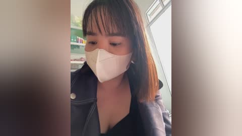 Video of an Asian woman with medium skin tone and straight, shoulder-length brown hair, wearing a black jacket over a black bra, and a white surgical mask, indoors with a blurry background.