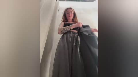 Media: Video of a fair-skinned woman with long, light brown hair, wearing a black crop top and loose, flowing gray skirt, standing in a narrow, dimly lit corridor with beige walls.