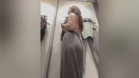 Media: Video of a woman with light skin and shoulder-length brown hair, wearing a gray dress, examining clothing items on a store rack in a brightly lit changing room.