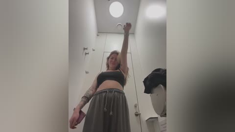 Media: Video of a tattooed woman in a bathroom, wearing a black sports bra and gray sweatpants, lifting a toilet brush, with a ceiling light and white walls in the background.