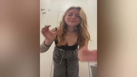 Media: Video of a young Caucasian woman with long, wavy blonde hair, wearing a black bra and gray sweatpants, making a peace sign in a white-walled room with a pink object in the background.