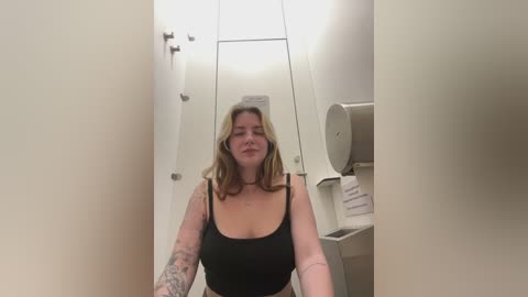 Media: Video of a middle-aged Caucasian woman with blonde hair, wearing a black tank top, standing in a narrow, brightly lit restroom with white walls and a roll of toilet paper dispenser.