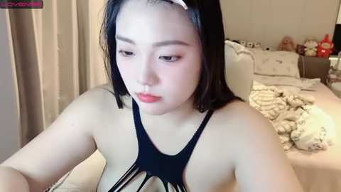 Media: Video of an Asian woman with fair skin, straight black hair, wearing a black halter top, sitting on a bed with a beige blanket, in a cozy bedroom with stuffed toys and a red bottle.