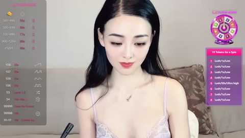 Media: Video of an Asian woman with long black hair, wearing a light pink lace bra, sitting on a beige sofa. Background shows a calendar and a \"Live\" stream banner.