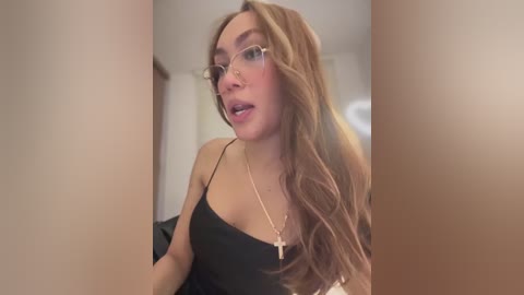 Media: Video of a light-skinned woman with long, wavy blonde hair and glasses, wearing a black spaghetti-strap top, standing indoors, looking slightly to the side.