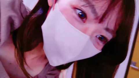 Media: Video of an Asian woman with long brown hair, wearing a white surgical mask, brown top, and visible red eye makeup, looking concerned, indoors.