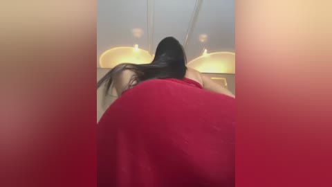 Media: Video of a woman with long black hair, leaning over a red couch, wearing a sleeveless red dress, in a modern, dimly lit room with soft lighting and beige walls.