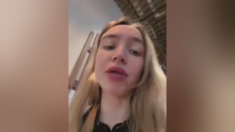 Video of a young Caucasian woman with long blonde hair, light skin, and pink lips, wearing a black top, taken from a low angle. Background shows a brick wall and a metallic object.