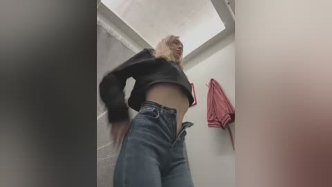 Media: Video of a blonde woman in a bathroom stall, wearing a black crop top and high-waisted jeans, with a red towel hanging on the wall.