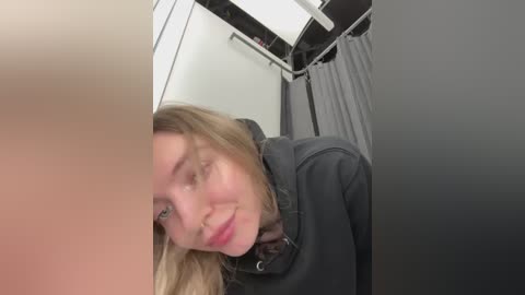 A video shows a smiling, fair-skinned, blonde woman with long hair wearing a gray jacket inside a dimly lit airplane bathroom. The background features a white wall, a gray curtain, and a white sink.