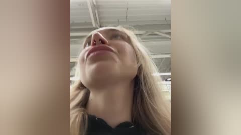 Video of a young woman with light skin, blonde hair, and fair complexion, looking up with a slight smile. She wears a black top and stands in an indoor setting with exposed white beams.